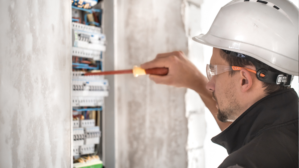 Elektryk w białym kasku montuje instalację elektryczną w rozdzielnicy przy użyciu śrubokręta.